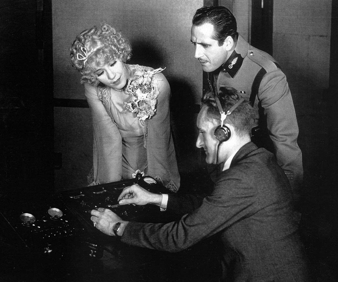 George Groves demonstrates the stage production mixer using headphones to John Miljan and Louise Fazenda in The Desert Song George Groves demonstrates the stage production mixer using headphones to John Miljan and Louise Fazenda in The Desert Song